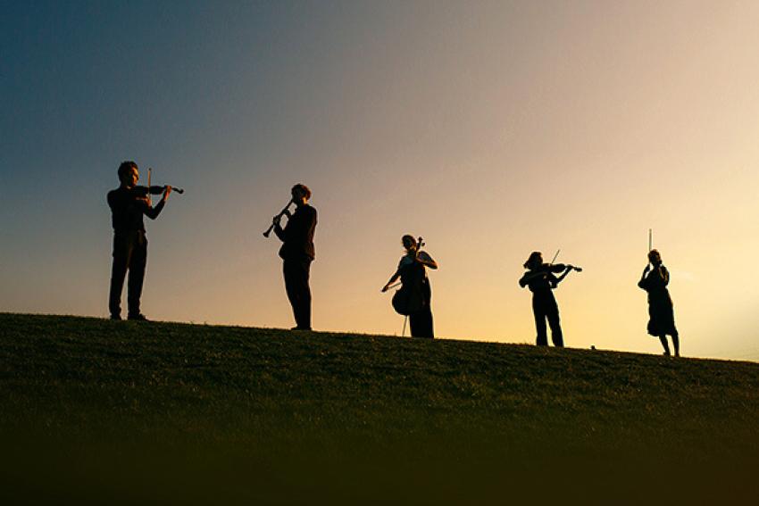 Silhouette style photograph of five musicians - string quartet and clarinet - on a hill at sunset
