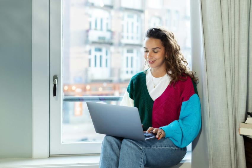 A smiling woman in a multi-colored jumper sitting by a window with a laptop