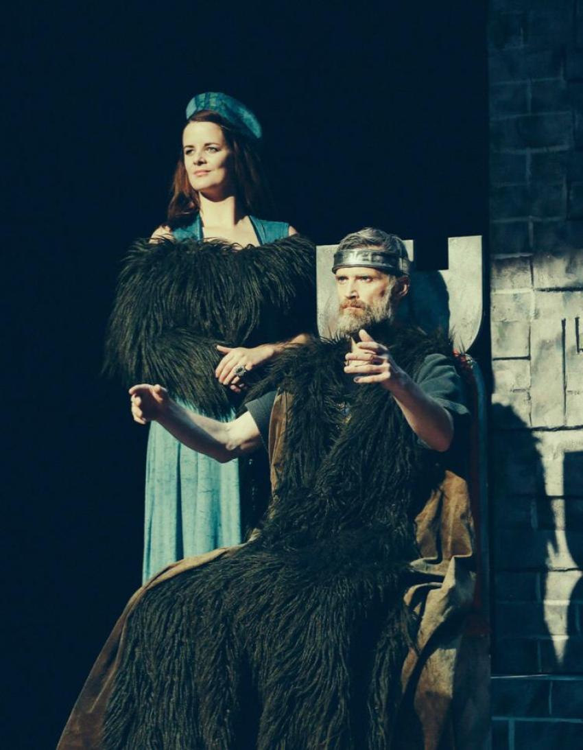 Macbeth sitting on a chair with Lady Macbeth standing beside him