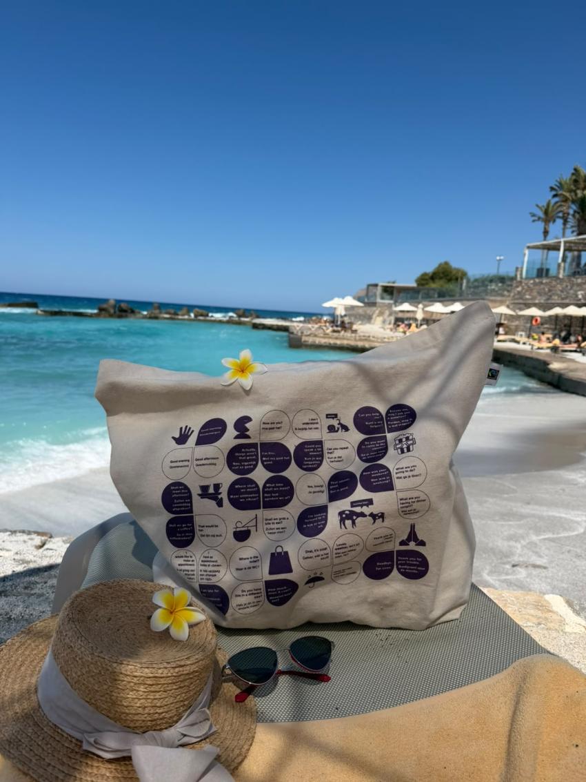 A Praat Nederlands Met Me beach tote bag on a beach with flowers, hat and sunglasses