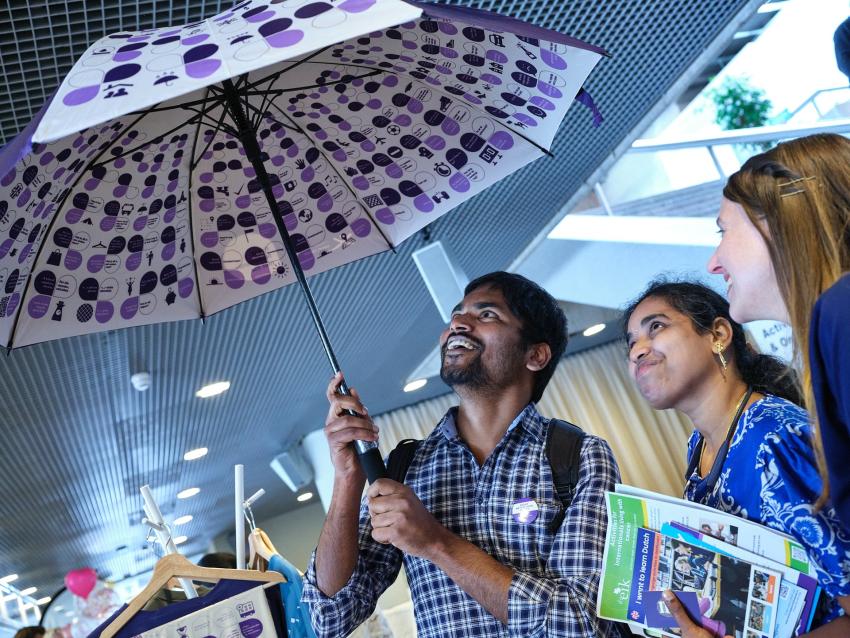 3 smiling people peeking under a Praat Nederlands Met Me umbrella