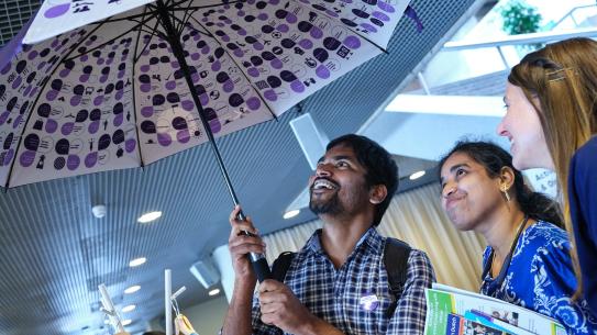 3 smiling people peeking under a Praat Nederlands Met Me umbrella, klik voor een vergroting