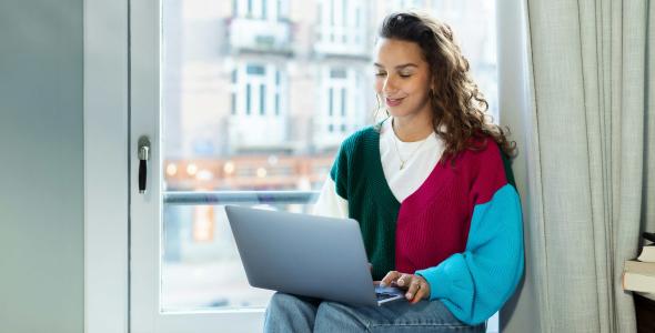 A smiling woman in a multi-colored jumper sitting by a window with a laptop