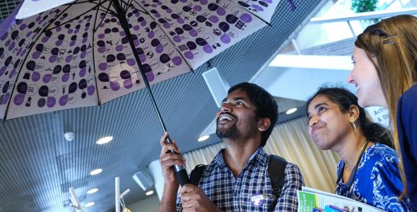 3 smiling people peeking under a Praat Nederlands Met Me umbrella